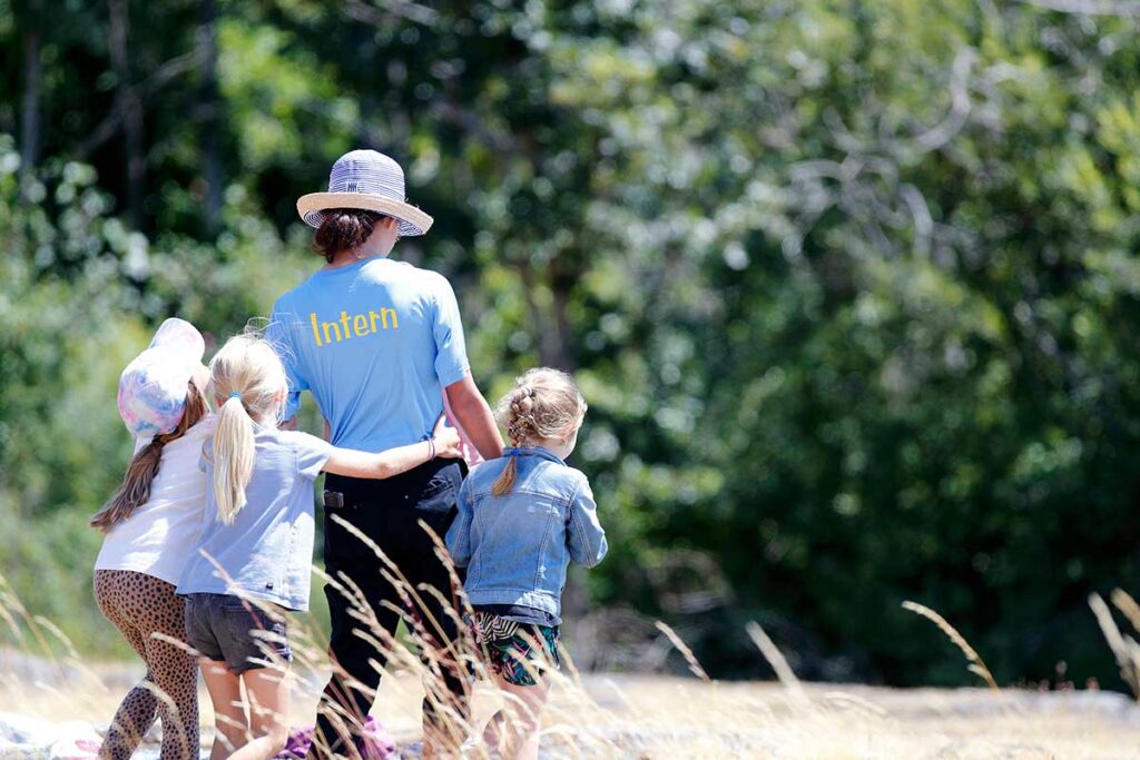 Three children and a camp leader walking with their back facing us outside in the sun.