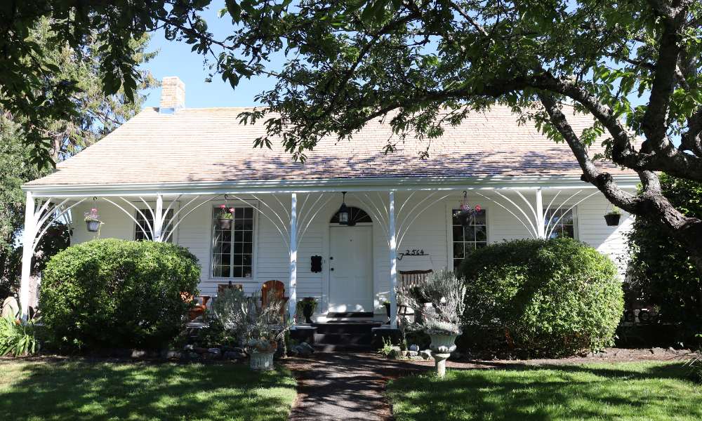 A white heritage house, single-level, in Oak Bay with bushes and trees.