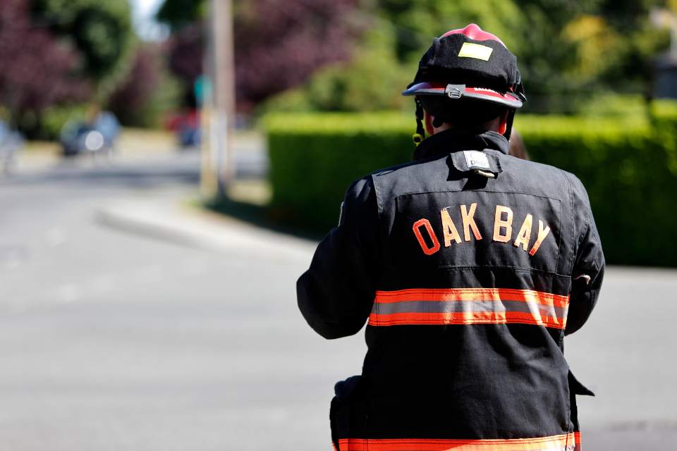 An Oak Bay Firefighter stands along a road, back facing the camera in full gear with the words "Oak Bay" on the back of their jacket.