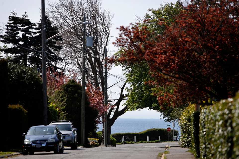 A road in Oak Bay with the ocean and in the background, trees lining the sidewalks and cars parked along the street.